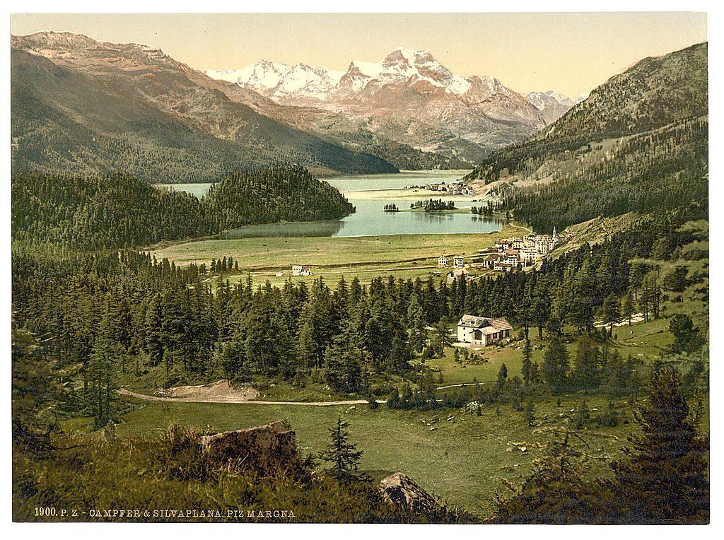 This image is a vintage colorized photograph depicting a serene and picturesque Alpine landscape. It captures the breathtaking views of mountains, forests, meadows, a lake with surrounding trees, several buildings that could be considered as houses or small towns, scattered in an orderly fashion around what seems to be a central body of water which looks like a lake. The foreground features dense greenery indicative of coniferous forestation typical of higher altitudes and the background is dominated by majestic snow-capped peaks. In the center distance lies another large expanse of water with several small islands or peninsulas, hinting at a larger river system beyond what's visible in this frame. The colors are muted greens, earthy browns, grays from rocky outcrops and mountain tops, punctuated by patches of white snow on distant peaks. A single caption reads '1900 P Z - CAMPFER & SILVA PLANA PZ MARGNA', suggesting the photograph's origin or its subjects' names associated with it during that time period in Upper Engadine region of Switzerland