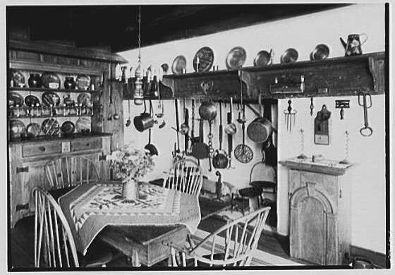 This black and white photograph depicts a traditional kitchen with an emphasis on vintage cooking utensils. Hanging pots, pans, plates, cups, and various other dishes are displayed in organized rows across the wall above a wooden dining table set for two people.

The room features exposed brickwork painted light gray underneath decorative tiles. A simple china cabinet houses additional white dinnerware to one side of the main area. The tabletop has an old-fashioned quilted runner with flower pot plants and a vase filled with flowers, which adds warmth to the space.

A wooden rocking chair is placed next to the table, while another stands at the end of it. Above the chairs hangs more kitchen tools such as ladles or spatulas. A vintage telephone sits on one side near an old-fashioned gas meter attached to the wall for domestic use and a small cabinet with doors in white paint.

The scene conveys a nostalgic atmosphere typical of early 20th-century life, suggesting that it could be part of Dr. A.C. Barnes' residence known as "Ker-Feal" located in Chester Springs, Pennsylvania dated August 20,1942.