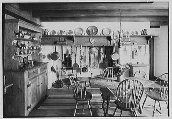 This black and white photograph depicts a traditional kitchen and dining area, likely from the early to mid-20th century. The setting appears homely with wooden furniture including chairs around a table adorned with a patterned tablecloth. Above them on shelves are various items such as plates, cups, utensils, pots, pans, ladles hanging above it all is an array of kitchen tools and dishes arranged in orderly fashion against the wall, giving off a rustic feel to this well-used space.