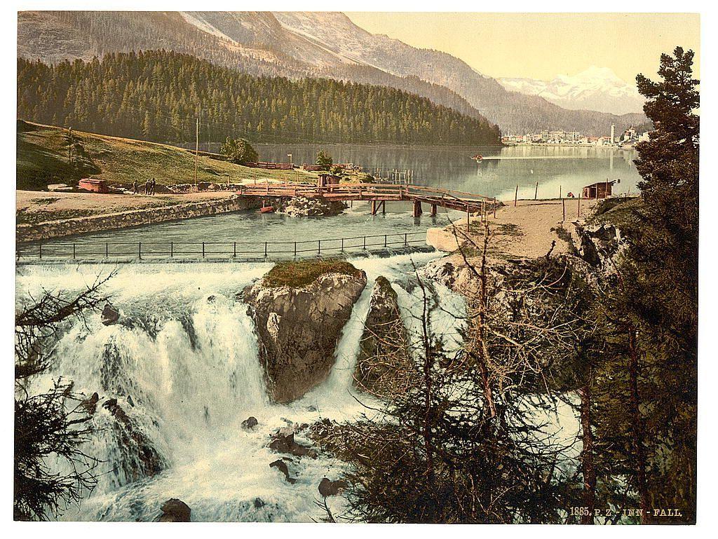 The image depicts a picturesque landscape featuring a waterfall cascading over rocks into the river. The surrounding area is lush with trees and greenery, leading up to mountainous terrain in the background which appears hazy possibly due to distance or atmospheric conditions. A wooden bridge extends across part of the scene connecting different parts of an industrial-looking facility on one side with buildings visible along a lakeside town on the other side. The overall color palette is muted with greens from vegetation, grays and browns for rocks and structures, as well as blues in the waterways reflecting the sky above. A copyright notice at bottom right indicates it's likely part of an older collection or archive.