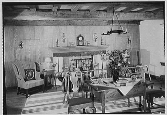 The image depicts a vintage living room setting with wood-paneled walls and ceiling, featuring rustic charm. A prominent fireplace serves as the focal point in the center of the space, adorned with various decorative items including framed pictures above it and an assortment on its mantle shelf. To the left stands a tall chair upholstered in striped fabric, accompanied by another armchair covered in patterned material near the window. The right side shows a wooden dining table set up for use, furnished with chairs around it and topped with vases of flowers, books, and other items suggesting daily life activities. A variety of lighting fixtures hangs from above – including wall-mounted lamps, an overhead chandelier, and pendant lights that cast shadows across the room's textured surfaces. The floor is partially covered by a striped area rug beneath the dining table. Overall, this image captures a cozy domestic scene typical of mid-20th-century American living quarters.