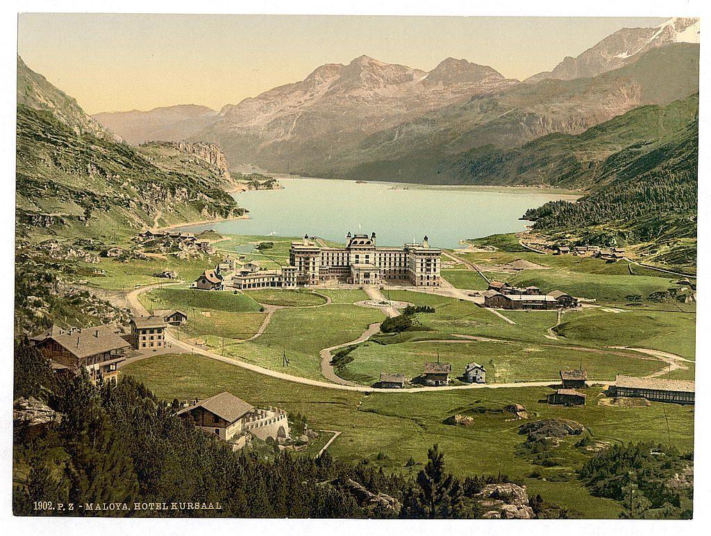 This image is a vintage postcard depicting an aerial view of the Maloya Hotel Kursaal in Switzerland, dated around 1902. The grand hotel stands prominently at its center amidst lush greenery and surrounded by mountains with peaks that fade into misty haze in the background. A serene lake stretches out to one side of the expansive grounds where smaller wooden chalets are scattered across the landscape. The coloration suggests it's an old postcard, as indicated by discolorations and slight creases on its surface. This picture provides a glimpse into early 20th-century Swiss tourism when such hotels were major attractions for European visitors seeking to enjoy mountain retreats.