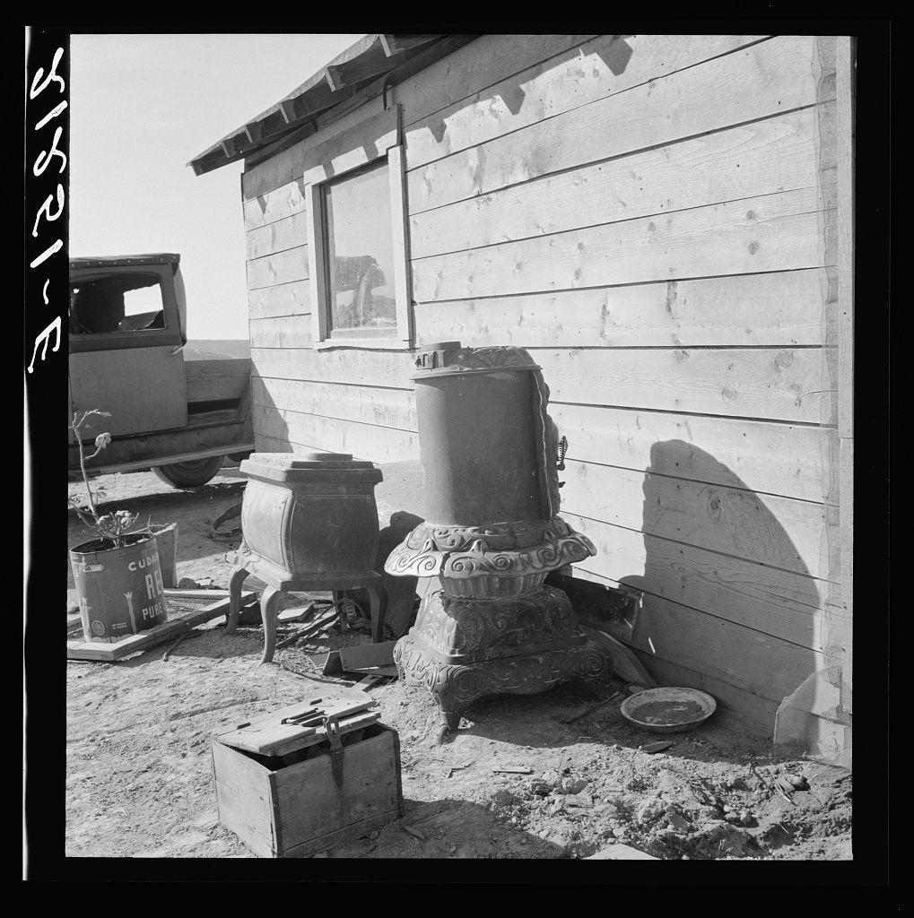 The image displays an outdoor setting with a wooden cabin and several pieces of old stove equipment in the foreground. The scene is captured in black and white, emphasizing textures and contrasts rather than colors. There's one large metal stove on its base, likely made from cast iron or steel, suggesting it has been used for heating purposes. Next to this main item are what appear to be parts disassembled from another similar stove: a smaller drum-shaped object with ornate detailing at the top, and two more cylindrical components that may have been part of the stove's base structure.

These stoves show signs of wear and aging; rust spots suggest exposure to moisture or weather elements. The surrounding ground is uneven and covered in dirt, which adds to the rustic ambiance of the scene. In addition to these items are a couple of other objects: an old wooden box with its lid off, lying on the ground next to one stove part, possibly used for storage; a metal pan that could have been used as a cooking pot or perhaps even a frying pan rests near another object.

The cabin in the background has vertical siding typical of rural dwellings built during early 20th-century America. The structure is simple with no visible windows on this side and lacks modern amenities, further suggesting an older setting possibly before electric heating became common.

In summary, this photograph  [...]