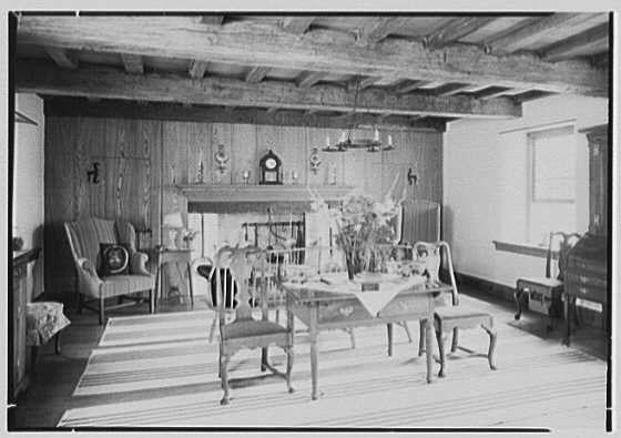 The image depicts a vintage-style living room with wooden paneling on the walls and exposed beams overhead. The focal point of the room is an old-fashioned dining table set for four, surrounded by matching chairs with curved backrests. On top of the table sits a vase filled with flowers, adding a touch of color to the otherwise monochromatic scene.

To one side of the room, there's a cozy seating area featuring two armchairs and a sofa, all upholstered in striped fabric that complements the overall aesthetic. A fireplace serves as the central heating source for this snug space, while various decorative items such as lamps, vases, and framed pictures adorn the mantel above.

The flooring consists of wooden planks laid out horizontally across the room, with contrasting stripes on a rug beneath the dining table to create visual interest. The natural light streaming in through a window illuminates the interior, highlighting the textures and craftsmanship of this well-preserved historical living space.