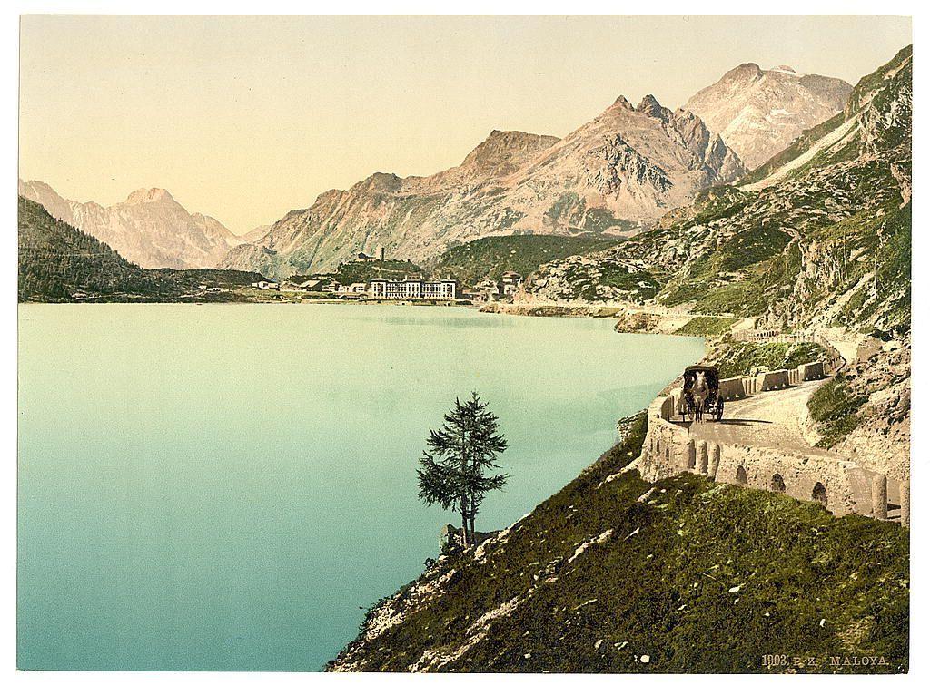 The image depicts a picturesque landscape featuring a serene turquoise lake nestled amidst majestic mountains. A stone road winds along the side of the mountain, with a horse-drawn carriage traveling on it, adding to the sense of tranquility and timelessness. In the distance, an elaborate building resembling a grand hotel or spa overlooks the water, complementing the natural beauty surrounding it. The sky is clear and soft-hued, suggesting early morning or late afternoon light. A solitary tree stands at the edge of the road, near which a figure appears to be seated on rocks, perhaps enjoying the breathtaking view. This idyllic scene captures an era where nature's splendor was preserved alongside human-made elegance in Switzerland during the 19th century.
