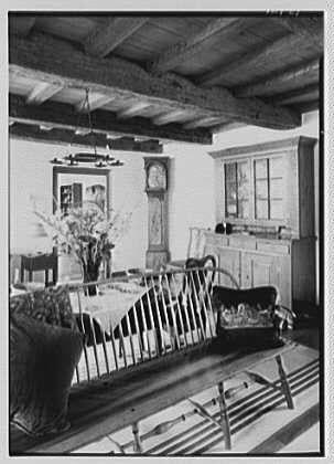 This black and white photograph shows a living room with various pieces of furniture, including chairs arranged around what appears to be a dining table. There is an old-fashioned sideboard against one wall, on top of which sits a large vase filled with flowers. A clock can also be seen in the corner near the ceiling beams that run across the room.
The photograph was taken as part of Dr. A.C. Barnes' residence located at Ker-Feal in Chester Springs, Pennsylvania in August 1942. The photo appears to have been preserved by Gottschol-Schleisner Inc., a company known for its historical photographs and collections from the early twentieth century.
The negative used for this photograph was captured on safety film with dimensions of 5x7 inches.
