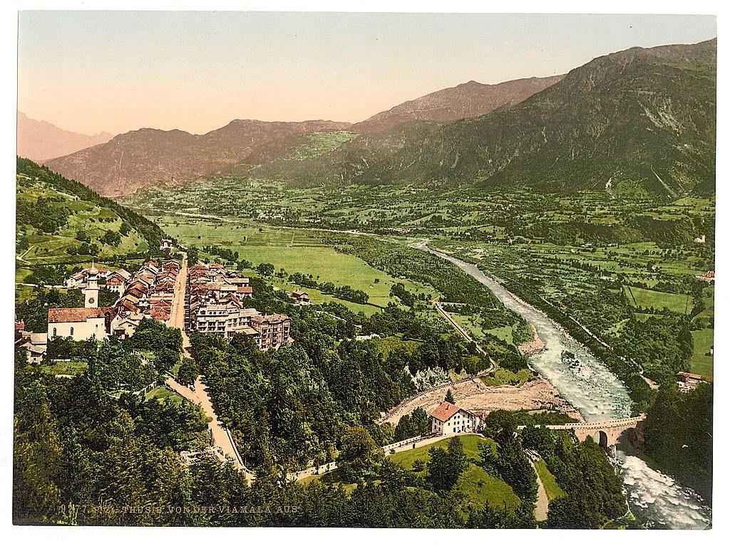 The image depicts a picturesque, rural landscape with rolling green hills and mountains in the background. A small town or village is nestled among these natural features, characterized by buildings with red roofs that suggest European architecture. The village is situated beside an expansive river flowing through its midst, which appears to have carved out winding paths around various terrains. Bridges cross over parts of the river within the vicinity of the settlement.

The surrounding fields and meadows are lush and green, indicative of fertile farmland possibly used for agriculture or grazing livestock. Paths or roads seem to connect different areas in the village and lead through some forested patches on either side. The overall scene is serene with a gentle blend of human habitation and untouched natural beauty, typical of rural settings within mountainous regions.

The colors are rich but muted, suggesting that this photograph might be quite old, possibly from around late 19th to early 20th century based on the image quality and coloration. There is no visible modern infrastructure or technology in sight, reinforcing its historical context as a snapshot of rural life during an earlier era.

Incorporated into the landscape are several prominent structures such as church spires and possibly old mills or other communal buildings which hint at both religious community  [...]