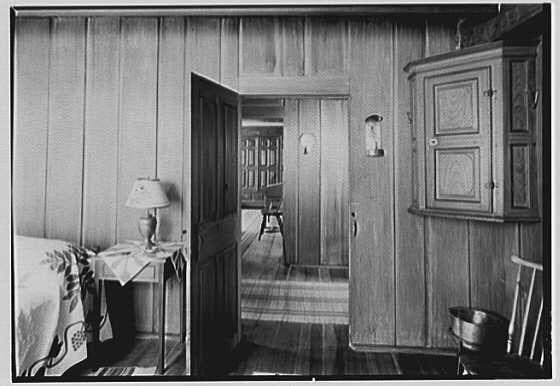 The image depicts a vintage black and white photograph of an interior room with wooden paneling. There's a doorway leading into another part of the house, revealing more wood-paneled walls and a chair placed in front of it.

In the foreground on the left side is a nightstand or small table beside which there appears to be a draped cloth item possibly serving as bedding material, indicating that this could be near a sleeping area. A lamp with a patterned shade sits atop the piece of furniture, providing light for reading or nighttime use. 

The room features wooden floors and has various elements such as a tall cabinet on wheels in what seems like an entryway to another space, possibly used historically for medical purposes given its sturdy construction.

Further back through the doorway is a larger area with chairs arranged around tables, suggesting it could be a common living or dining space. The overall ambiance of this setting conveys simplicity and functionality typical of early 20th-century rural American homes.