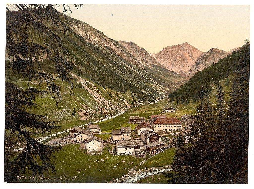 The image depicts a picturesque landscape featuring a small mountain village nestled within verdant slopes. The foreground is dominated by rustic buildings, including chalets and farmhouses with sloped roofs indicative of alpine architecture. A meandering stream cuts through the scene, reflecting sunlight on its rocky bed as it flows towards an out-of-sight point in front of the villas.

The middle ground reveals lush greenery leading up to a river valley that winds into the distance. The verdant landscape suggests late spring or summer with ample foliage and absence of snow. Towering mountains form a majestic backdrop, their slopes adorned with dense forests at lower elevations transitioning to barren rocky outcrops higher up. Among these peaks stands a prominent peak in the background, its gentle slopes leading down towards the village.

The sky is clear but tinged with hues that suggest either dawn or dusk; it's not overly bright yet carries enough light for visibility of details across the valley and mountainous terrain. The colors are naturalistic, consisting primarily of greens from trees and grasses, browns and grays from buildings and rocks, as well as touches of blue in the sky.

The photograph bears a caption at its bottom edge: '9278.P.Z.-SCARL,' which likely indicates that it's part of a collection or series. The overall composition exudes tranquility and beau [...]