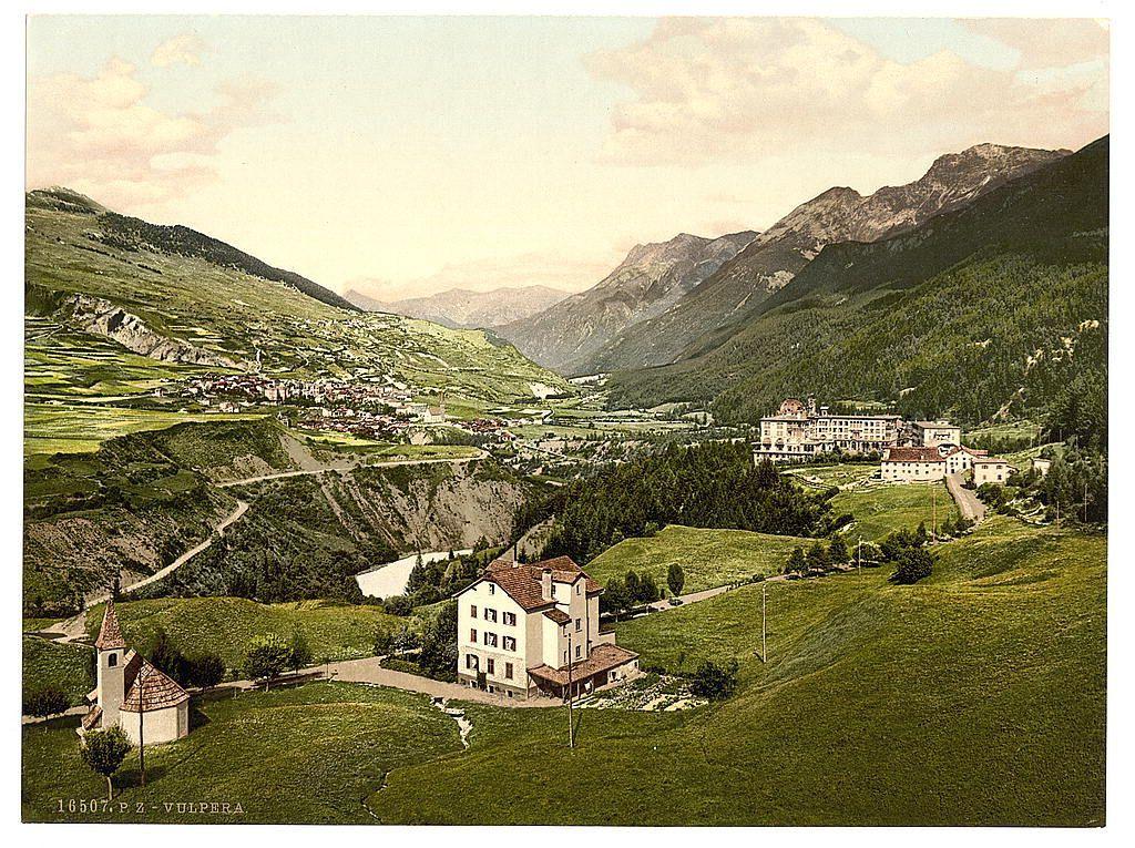 This image depicts a scenic landscape with lush green hills, dotted houses and buildings nestled in the valley. In the foreground stands an old church-like structure with a spire on its roof, situated next to another building with red-tiled roofing. Further back, a larger mansion or hotel is visible atop rolling fields, surrounded by several more structures that appear as part of what might be a resort complex. The backdrop features majestic mountains rising steeply from the valley floor, their peaks partly obscured by clouds. A winding road snakes through the hillsides leading up to and away from this central building cluster.

The photo has an aged quality with sepia tones indicative of early color printing techniques like photolithography or chromolithography used during the late 19th century in places such as Switzerland, Germany, Austria-Hungary (which included present-day Slovakia), Russia, France and Eastern Europe. The image is labeled "Vulpera" at its bottom left corner, suggesting a specific location within this region of interest.