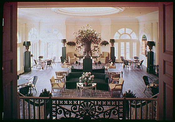 The image depicts a spacious, well-lit sunroom with high ceilings and large windows that allow natural light to flood the space. The room is furnished in an elegant style featuring several pieces of furniture such as sofas, chairs, tables, and armchairs arranged around a central area rug.

At one end of the room stands a tall tree-like plant housed in a dark pot, adding a touch of nature to the decor. Several potted plants are strategically placed throughout the space, enhancing its inviting atmosphere. The flooring appears to be made of polished wood or possibly marble tiles that reflect some light and add to the luxurious ambiance.

The walls are painted in a soft beige color with decorative moldings around the windows and door frames adding an element of sophistication. Accent lighting fixtures on tall slender stands contribute to the room's warm, inviting glow. The sunroom is enclosed by dark wooden railings adorned with intricate ironwork that allows for unobstructed views.

Overall, this image portrays a classic example of mid-20th-century interior design featuring both comfort and style in an elegant setting.
