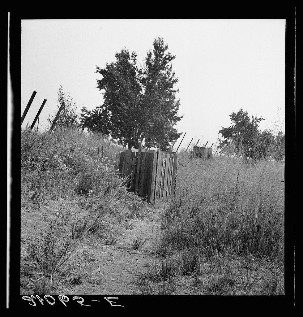 The image is a black and white photograph depicting an outdoor rural scene. It shows overgrown grass, tall trees in the background, and a wooden fence with vertical posts and horizontal rails that appears to be worn down or weathered. The ground seems dry with sparse vegetation. There are several broken pieces of metal wire scattered on either side of the pathway created by foot traffic through this area. In the foreground, there is some handwritten text which reads "10P2-E" at a diagonal angle across it in capital letters and numbers that could be part of an inventory or document reference system related to hop pickers' camps as indicated by the caption provided. The environment suggests neglect but still maintains a sense of rural tranquility with no human presence visible within this snapshot captured moment.