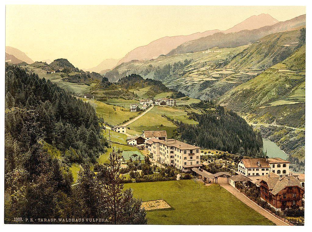 The image depicts a picturesque rural landscape with rolling hills and lush greenery. Nestled among the slopes are several buildings, including what appears to be a hotel or guesthouse complex in the foreground on the left side of the frame. The architecture is traditional European, possibly indicative of an alpine region given its style and setting.

A winding road cuts through the village, leading toward other structures scattered along the hillside paths. A body of water, likely a river, meanders from the background into the valley where these buildings are situated. Beyond the immediate landscape, mountains with gentle slopes rise in the distance under a clear sky tinged with warm hues.

The overall atmosphere is serene and idyllic, suggesting an untouched natural setting ideal for leisure or travel during that era. The colors in the image have muted tones, typical of colorized postcards from late 19th to early 20th-century Europe.