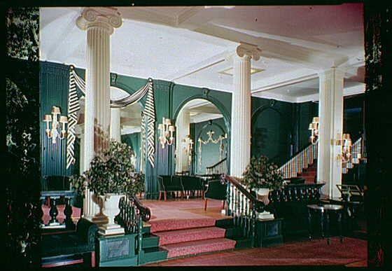 The image shows an elegant ballroom with a grand staircase at one end, decorated in dark green and red tones. The walls are painted richly colored deep greens and reds, accented by white pillars that line the room. A set of ornate chandeliers hang from the ceiling, casting a warm glow over the space. On either side of the entryway, tall lamps flank large potted plants with lush foliage on top, adding to the opulent atmosphere.

A staircase with red carpeting and wooden balusters leads up to what appears to be another level or area beyond the ballroom's reach. The room is furnished with dark wood chairs arranged around small tables where guests might sit for conversation or refreshments during events held here.

The overall ambiance of this space exudes sophistication, historical charm, and a sense of grandeur from an era long past, likely dating back to at least mid-20th century as suggested by the caption indicating it was taken in 1946. The image captures not just the physical details but also hints at its use for hosting social gatherings or events within this historic Virginia landmark.