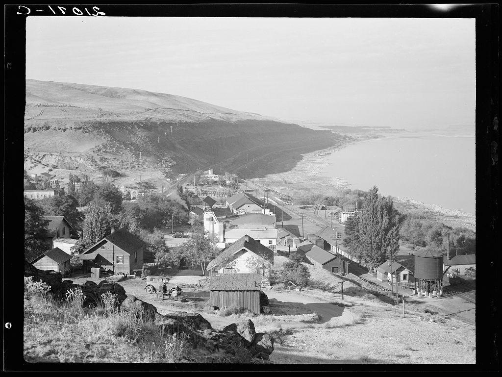 This black and white photograph captures a rural town situated on the banks of the Columbia River in Oregon. The perspective is elevated, providing an overview of several modest homes with varying architectural styles nestled among trees and open fields. A prominent water tower stands out towards the right side, while various other structures such as barns, sheds, and silos dot the landscape indicative of a farming community.

The backdrop features rolling hills leading down to the river's edge where sparse human presence is visible along the shoreline. The overcast sky suggests it could be early morning or late afternoon when shadows are elongated. This image appears historical in nature with its grainy texture, which might hint at an older era of photography before color film became prevalent.

The label "O-Trois" printed across one corner adds to this vintage feel and likely refers either metaphorically to the town itself being a 'third place' or perhaps is part of some coded information. The inclusion of such markings within black-and-white documentary-style photographs was common during that period, where photographers often added labels for context or personal annotations.

Additional details include an old tractor with its plow attached in front of one house and utility poles stretching across the scene signifying connectivity to a broader network. Overall, this im [...]