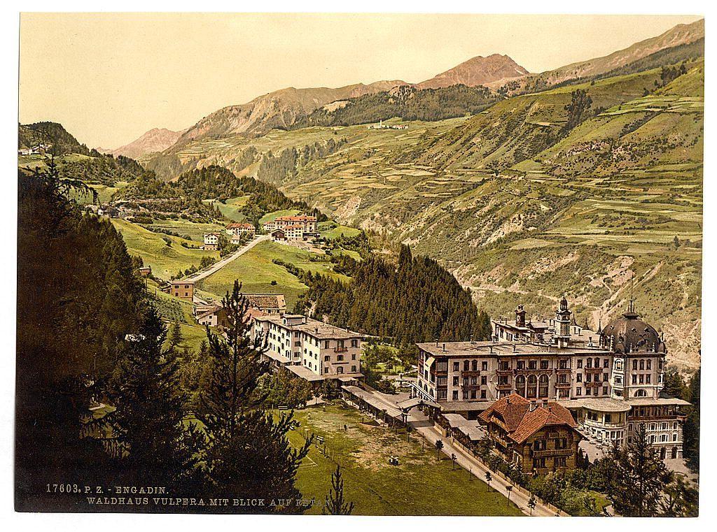 The image depicts a picturesque mountainous landscape, likely taken between the late 19th and early 20th century. It shows terraced fields on rolling hillsides leading up to dense forested areas at higher elevations. Nestled in this rural setting is an elaborate building complex with multiple wings and towers characteristic of historical or significant architecture from that era. The structure's design includes numerous windows, balconies, and ornate facades, suggesting it may have been a notable establishment such as a manor house, hotel, or administrative center.

Surrounding the main building are smaller structures, likely residential houses with traditional European architectural features like pitched roofs, wood siding, and small gardens. A meandering road snakes through the settlement, connecting various buildings and leading towards distant peaks partially obscured by mist. The warm hues of brown, green, and yellow dominate the scene, conveying a sense of tranquility and pastoral beauty.

The image bears text at its base indicating it is an old postcard or print from Engadin, Switzerland, specifically referencing Vulpera (likely located in Fetan) within Grisons department between 1890-1900. The inclusion of such information suggests that this area was a point of interest for visitors during the time period mentioned, potentially due to its scenic beauty and histori [...]
