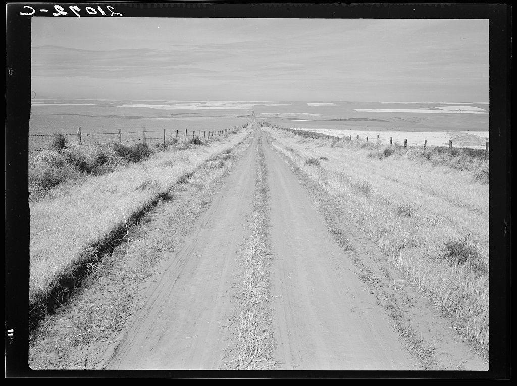 A black-and-white photograph depicts a dirt road leading towards the horizon in an open landscape. The terrain is flat and appears arid, with dry grasses on either side of the unpaved path that shows tire tracks indicating frequent use by vehicles. A fence made up of wooden posts connected by wire stretches along both sides of the road from left to right, marking a boundary or enclosure for land beyond it. In the distance, expansive fields recede into a hazy horizon where some areas are flat and others have slightly undulating terrain. The sky is overcast with clouds diffusing sunlight across the scene. Text overlay in white at the top of the image reads "3-spoils," possibly indicating a location or reference associated with this photograph.