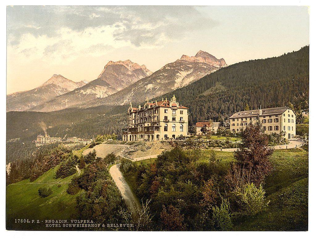 This image depicts a picturesque landscape featuring an elegant hotel nestled among rolling hills and towering mountains. The hotel, prominently situated in the foreground on lush green grounds, has multiple stories with decorative architectural elements such as ornate gables, dormer windows, and chimneys. Surrounding it is verdant foliage, creating a serene atmosphere. A winding road leads up to the hotel from the bottom left corner of the image.

In the background, majestic mountain peaks rise against a partly cloudy sky, adding depth and grandeur to the scene. The overall setting suggests an idyllic location in Switzerland's Grisons region during late 19th or early 20th century when such hotels were popular destinations for tourists seeking leisurely getaways.

The photo appears aged with some discoloration and wear indicative of its historical significance, providing a glimpse into the past charm and allure of Swiss alpine hospitality.