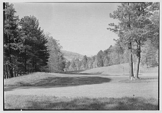 The image depicts a serene and expansive outdoor setting, seemingly captured during the mid-20th century judging by its black-and-white composition. It is dominated by an open field with neatly trimmed grass leading towards what appears to be a golf course fairway or path bending around it.

On either side of this central pathway are tall trees that stand as sentinels along the landscape, their foliage varying in shades from light to dark green suggesting different species and perhaps indicating changes in season. The leaves on these trees exhibit natural variability - some are fully leafed out while others have fewer or none at all, which could point towards a transition period between seasons.

In the background rises a gentle hillside with more sporadic foliage, leading up to what seems like dense woods or forest that extends beyond into the distance. The sky is clear and devoid of any clouds, suggesting fair weather conditions during the time this photograph was taken on May 15th in Hot Springs, Virginia as noted by the caption.

The absence of human figures or animals brings a sense of tranquility to the scene; it's an untouched moment frozen in time with nature's calm prevailing over potential activity associated with golf. The setting is peaceful and unspoiled which may indicate that this area was not heavily trafficked at the time, offering a glimpse into less imp [...]