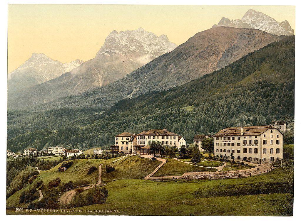 This image depicts a picturesque landscape featuring traditional European architecture nestled amidst rolling hills and towering mountains. The buildings are styled in a manner indicative of the late 19th century, with their symmetrical facades and classic window design suggesting an Alpine region. A series of white houses or villas form a cluster on elevated grounds, surrounded by greenery that suggests springtime growth. To the right stands a larger building named "Hotel Kulm," which indicates its use as an inn or lodging establishment.

The backdrop is dominated by majestic peaks blanketed in patches of snow and dense coniferous forests covering lower elevations, suggesting varied altitude zones within this mountain range. The overall palette consists of earthy greens and browns for the foreground vegetation and slopes, while the mountains display hues ranging from white to gray due to their snowy caps.

A dirt road winds through the scene, leading visitors on a journey towards or away from these stately structures. A small wooden structure stands out near one building's entrance, possibly serving as an information booth or gatehouse for guests arriving by carriage or footpath. The image is labeled with text at the bottom: "1908 P.Z.VULPERA., PIZ GI.JON., Piz LISCHANNA," indicating it captures a locale in the Engadine region of Switzerland, specifically mentioning Vulp [...]