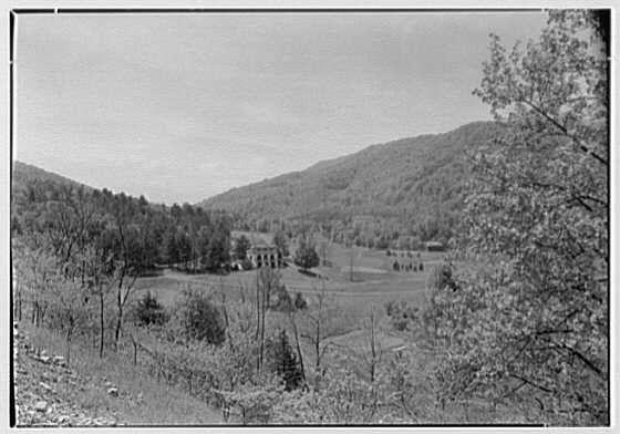 This black and white photograph depicts a scenic, rural landscape with rolling hills and sparse tree coverage. In the center of the image stands an old building that appears to be a clubhouse or estate; it has a prominent entranceway flanked by columns. The structure is surrounded by open fields and patches of cultivated land, suggesting agricultural activity in this area.

The foreground shows uneven terrain with some exposed soil and bare ground areas, possibly indicating recent clearing for farming purposes. Several young trees are visible throughout the landscape, hinting at reforestation efforts or early growth stages after a previous disturbance to the environment. The absence of people and modern infrastructure emphasizes the tranquil nature of this location.

The sky is clear but devoid of clouds, suggesting fair weather conditions during the time when the photograph was taken on May 15, 1946. Overall, the image captures a moment in history where rural America seems to have been preserved from rapid industrialization and urban sprawl. The source indicates that this photo belongs to Gottschol-Schleisner Inc., which is likely an archival company or photography studio responsible for documenting historical sites such as Hot Springs' Homestead resort, Cascades clubhouse.

This serene snapshot of the past offers a glimpse into America's bucolic heritage and serves as a [...]