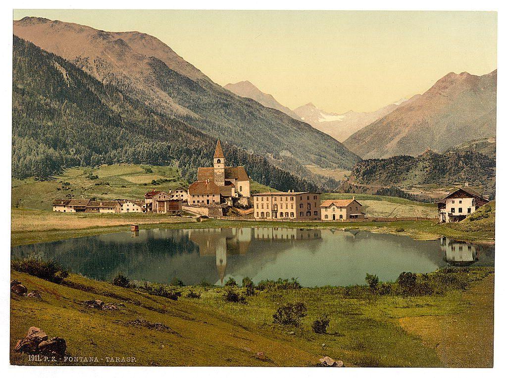 The image depicts a picturesque landscape with a quaint village nestled in the valley below towering mountains. The central focus is an old, stone church featuring a prominent bell tower and surrounding buildings that appear to be residential or possibly part of a monastery complex. To its right stands another substantial building with multiple stories, exhibiting features consistent with European architecture from past centuries.

In front of these structures lies a large pond reflecting the village's silhouette on calm waters, which adds symmetry to the composition. The foreground showcases vibrant greenery and scattered boulders near the water's edge, indicating an undisturbed natural setting. Rolling hills roll into the distance where more rustic buildings dot the terrain, and snow-capped peaks loom in the far background under a hazy sky.

The photograph is marked with "1911.PZ" at its lower right corner, suggesting it was printed or published around that year. The inscription reads "FONTANA-TARASP," pointing to Tarasp village within Switzerland's Graubünden region (Grisons). This scenic view represents the beauty and tranquility of rural areas in this part of Europe during an era where such locales were popular subjects for postcard photography, offering a glimpse into both historical architecture and natural scenery.