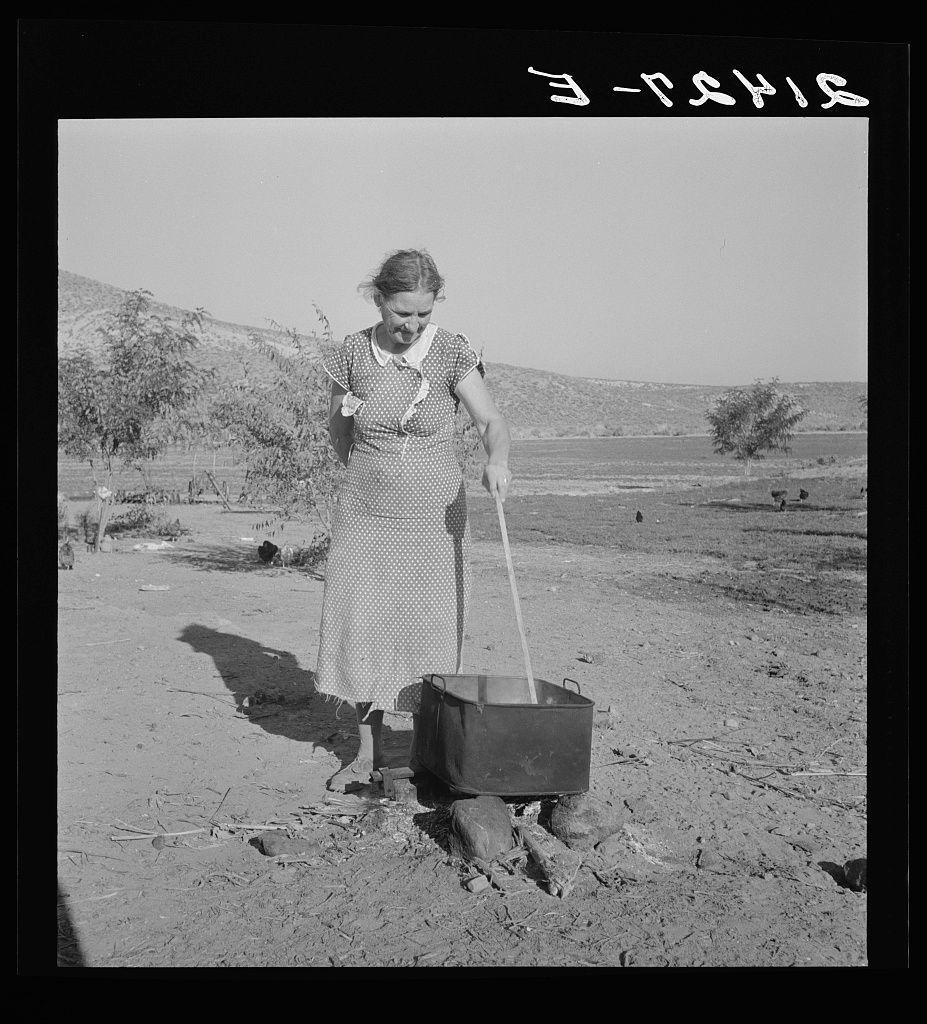 The image depicts a black and white photograph of an older woman standing outdoors with her legs partially buried in the ground, using what appears to be a shovel or pitchfork to stir a metal pot on top. She is wearing a polka-dotted dress with puffed sleeves and has short hair that reaches just past her shoulders. The background shows a barren landscape with sparse vegetation and trees under a clear sky.

The photo includes text at the bottom right corner, which reads "E-T-S-H-I" in reversed letters due to the image being oriented upside down. Additionally, there's handwritten white lettering on the top left side of the photograph that appears to be written by hand but is difficult to decipher completely from this angle and resolution.

The overall atmosphere suggests a rural or agricultural setting, with an emphasis on manual labor for food preparation in outdoor conditions. The image may evoke themes related to historical farming practices, daily life during early 20th-century America, specifically focusing on the working class' struggle against harsh living conditions through perseverance and resilience.