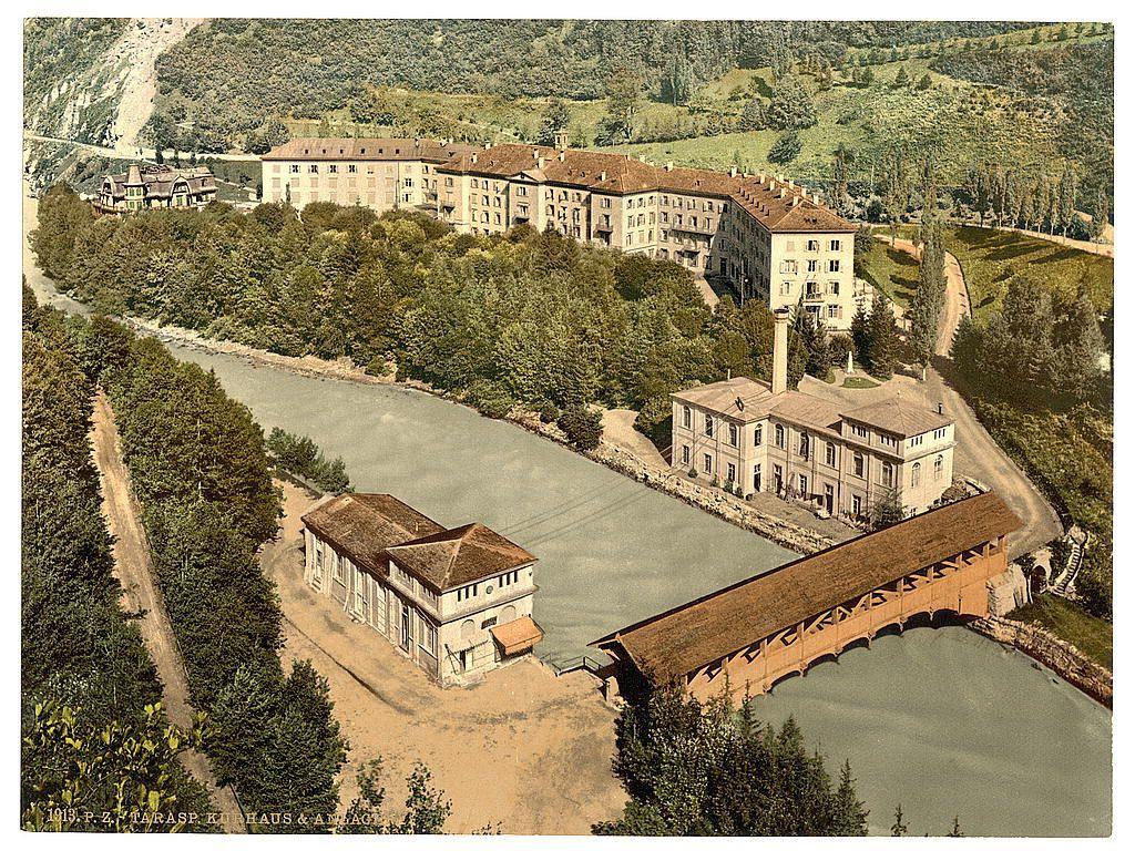 The image depicts an aerial view of a European village or town, possibly from the late 19th century. It shows several multi-story buildings with sloped roofs and large windows, situated on hilly terrain surrounded by dense forests. A river meanders through the landscape, partially obscured by greenery, winding its way around the settlements.

A prominent wooden bridge spans across a stretch of water in the foreground, indicating human intervention to connect different parts of the town or valley. The buildings exhibit features typical of European architecture from that era, with some displaying ornate facades and others more functional designs suited for communal purposes such as schools or administrative offices.

The colors are muted earth tones—greens of trees, browns and reds of rooftops and structures—with a sepia-like tint suggesting an old photograph. The caption indicates the location is Lower Engadine in Tarasp, Kurhaus Grisons (Grisons), Switzerland, dated approximately between 1890 and 1900.

Given these details, it's clear this image captures historical architecture within a rural Swiss landscape during its turn-of-the-century past.
