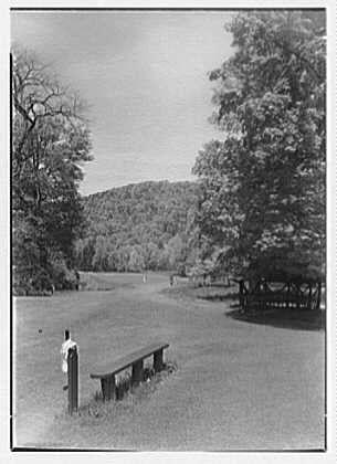 The image depicts a tranquil outdoor scene, likely from the early 20th century given its black and white composition. In the foreground, there's an open grassy field with neatly trimmed edges extending into the distance where it meets rolling hills or mountains covered in dense trees.
A solitary wooden bench sits on one side of the path, suggesting a resting spot for golfers enjoying the course. To the left, near this bench stands a tall figure wearing period attire - likely indicative of either a golfer or an attendant from that era.

The sky is mostly clear with only slight cloud cover visible above. The absence of modern-day equipment and clothing styles indicates a historical context to this photograph capturing leisure activities in what appears to be the early days of golf as we know it today.