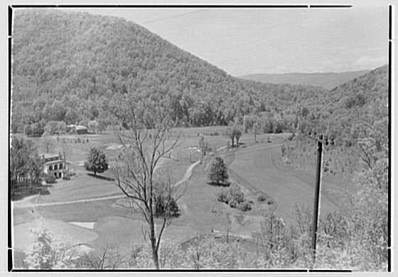 The image depicts a scenic, rural landscape with rolling hills and lush vegetation in the background. In the foreground, there is an expansive open field that appears to be part of a golf course or sports ground, characterized by patches of grassy areas and some scattered trees without leaves, suggesting it might be taken during autumn or winter.

A building resembling a clubhouse can be seen on one side of the frame, indicating human activity in this otherwise natural setting. A winding road cuts through the middle section of the image, adding depth to the scene as it snakes its way around the landscape.

The photograph is presented in black and white, which gives it a timeless quality or possibly indicates that the photo was taken several decades ago when color photography wasn't prevalent. The overall composition provides a tranquil ambiance, capturing both nature's beauty and signs of human leisure pursuits within this serene environment.