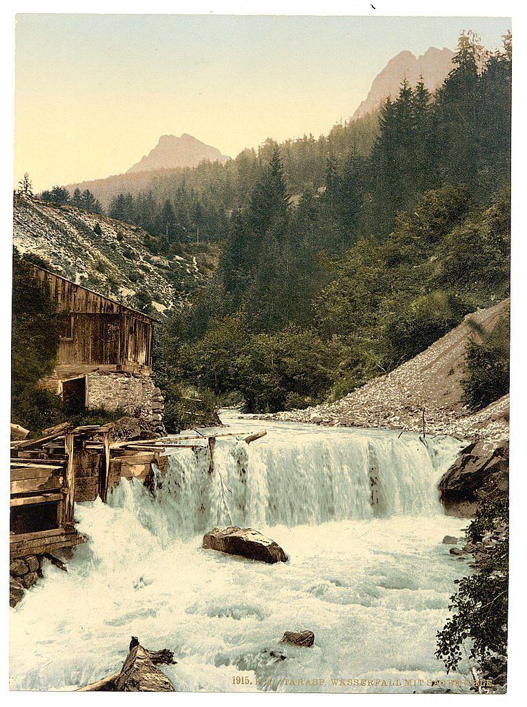 The image depicts a serene landscape featuring a waterfall cascading down rocks with force. In the foreground, there is a rustic wooden structure adjacent to the water flow, which appears aged and weathered by time. The surrounding environment showcases lush greenery indicative of forested terrain, with dense trees on both sides of the stream that create a natural corridor leading towards distant mountains in the background. These peaks are partially obscured by mist or cloud cover, adding an air of mystery to the scene.

The overall composition exudes tranquility and highlights the untouched beauty of nature. The colors are rich yet muted, emphasizing greens of vegetation against the browns of wooden structures and rocks amidst flowing white water.