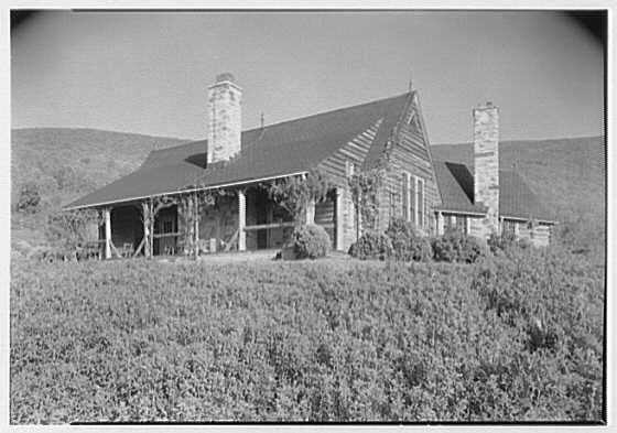 This black and white photograph depicts a rustic log cabin with two prominent chimneys on either side of the structure. The building features a steeply pitched roof, large windows covered by what appears to be curtains or drapes, and an expansive porch supported by thick wooden beams. Surrounding the cabin is dense vegetation that seems like wild grasses or brush.

The setting suggests it could be located in rural countryside, with rolling hills visible in the background under a clear sky. The overall scene evokes a sense of historical charm and simplicity associated with pioneer architecture.