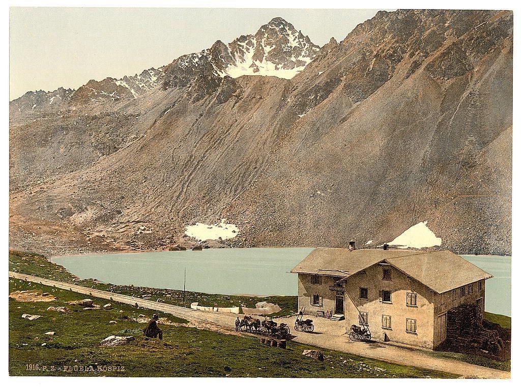 The image depicts a picturesque landscape featuring the Fluela Hospice in Grisons, Switzerland. This historic photograph captures an alpine scene with towering mountains blanketed by snow-capped peaks dominating the background against a clear sky.

In the foreground, there's a quaint stone building labeled "Fluela Hospiz," indicative of its function as a mountain hut or shelter for travelers. The architecture is simple yet sturdy, reflecting early 20th-century design sensibilities with large windows and exposed wooden beams.

The hospice appears to be nestled on lush green grassy slopes leading down towards the serene turquoise waters of a lake that occupies much of the middle ground in the image. A paved pathway or road runs alongside the building, suggesting it's accessible for visitors and possibly serves as a route connecting travelers through this remote region.

Several individuals are visible around the hospice: one person is seated on a rock near the water's edge, while others stand by horse-drawn carts loaded with supplies, indicating their use in transporting goods. The presence of these vehicles suggests that during its operation, it played an essential role for those traversing this challenging terrain.

Overall, the image exudes tranquility and charm, portraying Fluela Hospice as a serene retreat within Switzerland's picturesque landscape, capturing both natu [...]