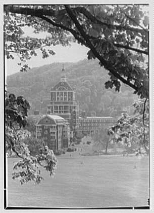 The image shows a black and white photograph of The Homestead, Hot Springs, Virginia. In the center stands an elegant Victorian-style building with ornate architectural details such as arched windows and intricate trim work on its facade. This structure is likely to be one of the main buildings in this historic hotel complex.
Behind it lies another large, classical-looking edifice featuring columns at its entrance and a series of balconies lining its upper levels. The surrounding landscape includes lush greenery with trees framing the foreground and midground views against a backdrop that suggests rolling hills or mountainous terrain shrouded by clouds or mist in the distance.
The overall ambiance conveyed is one rich with history, evoking an air of grandeur and opulence typical for such luxury resorts during its heyday. The photograph appears to capture this scene from across what could be considered today's pool area, providing a picturesque perspective that showcases both architectural beauty as well as natural surroundings harmoniously blending together.
The image is credited to Gottleosch-Schleisner, Inc., who likely provided professional photography services at the time of its creation. The negative appears to have dimensions 5x7 inches which indicates it was produced in a standard photographic format used for large-format negatives during this period.
Furthermore,  [...]
