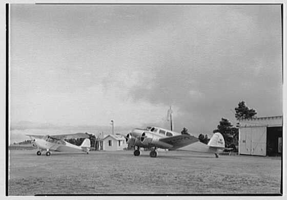 The image depicts a black and white photograph of several vintage propeller aircraft parked on what appears to be an airstrip. The planes are various models with different wing shapes, engine types, and seating configurations. In the background, there is a small building which could serve as an airport office or hangar, along with other infrastructure like light poles and possibly control towers in the distance.

The sky above is overcast, suggesting cloudy weather conditions during the time of photography. The aircraft are positioned at different angles relative to each other, indicating they may be prepared for various stages of flight operations such as takeoff, landing, or maintenance checks.

There's a watermark on this image that reads "Gottscho-Schleisner, Inc." which suggests it is part of their collection and likely taken during the period mentioned in the caption. The presence of such an aircraft photograph could signify its historical value for aviation history enthusiasts or to document early 20th-century air travel.

As a piece of archival material from May 15, 1946 at Hot Springs Airport, Virginia, this image provides insight into aviation's past and captures the essence of mid-20th century aircraft design.