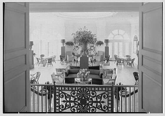 The image depicts a luxurious sunroom viewed from above, likely within The Homestead in Hot Springs, Virginia. It showcases an elegantly furnished space with dark wood chairs and tables arranged throughout the area. In the center stands a large potted plant surrounded by numerous small lamps placed on various surfaces. A decorative wrought-iron railing adds to the room's sophisticated ambiance.
The sunroom is illuminated through multiple windows, revealing additional furnishings like couches and more plants in the background. The overall atmosphere suggests an elegant and serene environment designed for leisurely activities or social gatherings during sunny days.