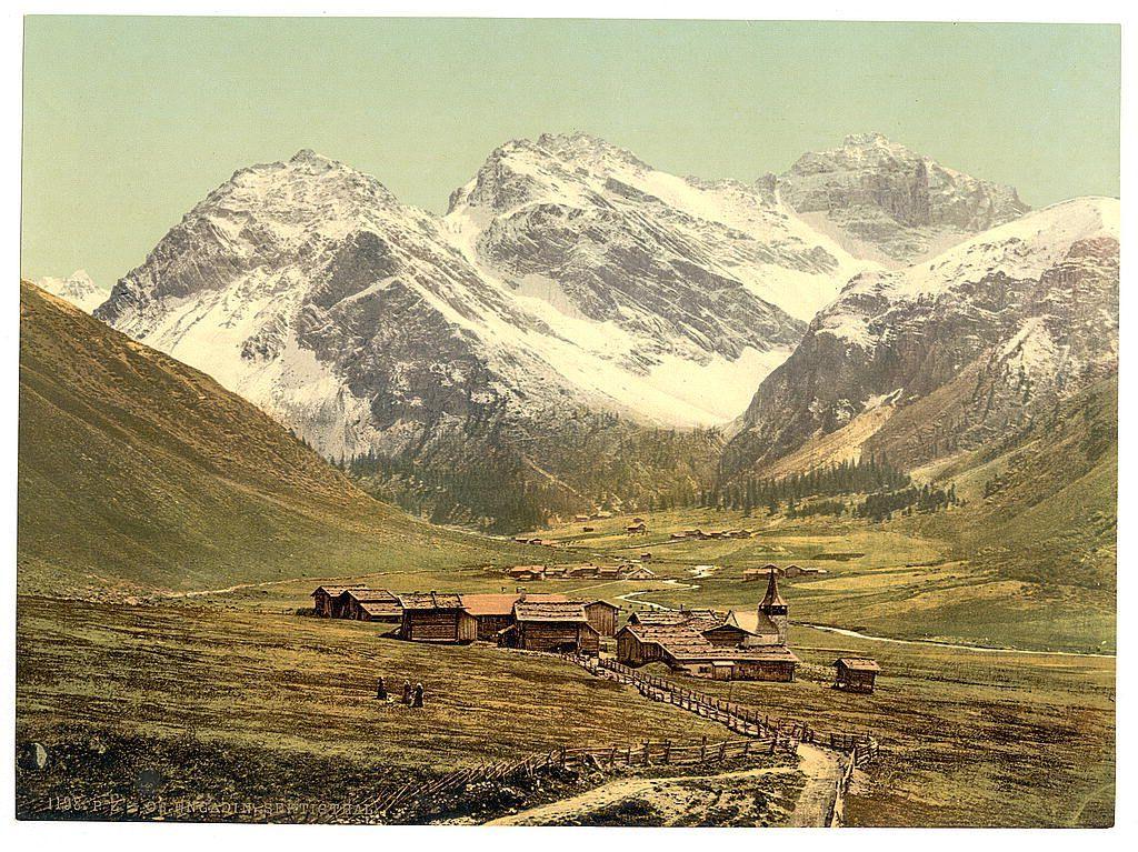 The image depicts a picturesque landscape dominated by towering snow-capped mountains that rise majestically against the sky. In the foreground, there is a quaint alpine village composed of several wooden chalet-style houses with sloping roofs and visible chimneys, suggesting warmth within. The structures are nestled in gently rolling fields dotted with patches of grass and sparse vegetation indicative of high-altitude conditions.

The settlement appears isolated yet harmoniously integrated into its environment, signifying the simplicity and self-sufficiency often associated with mountainous communities during that era. There's a sense of tranquility as no modern elements disrupt this natural setting.

A winding path or road cuts through the fields leading towards the village entrance from the bottom right corner, inviting viewers to imagine their own journey into this serene world. In the background, dense conifer forests add depth and contrast against the stark white snow on the mountainside, enhancing the overall dramatic beauty of the scene. The colors are rich yet muted with earthy tones dominating the palette.

The image is reminiscent of early colorized postcards or prints from that time period when photography was still in its infancy before full-color processing became widespread. It captures a moment frozen in history, offering viewers insight into life and land [...]