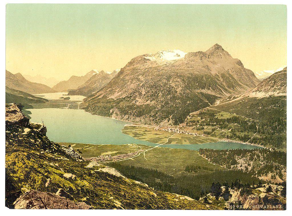 The image depicts a stunning natural landscape, showcasing the beauty and grandeur of mountainous terrain. In the foreground, steep slopes are adorned with patches of greenery, possibly grass or shrubs, indicative of alpine flora adapted to rocky conditions. The middle ground is dominated by a serene lake reflecting the sky above; its waters have shades of blue-green, contrasting against the earthy tones of surrounding land.

Beyond this tranquil body of water lies a valley dotted with clusters of buildings and cultivated fields, suggesting human habitation nestled within nature's embrace. These structures appear small from this high vantage point but indicate an active community living in harmony with their environment.

The background is dominated by majestic mountains rising sharply against the sky, some peaks blanketed partially or fully in snow, highlighting differences in altitude or seasonal variations. The overall palette of colors ranges from vibrant greens and yellows to deeper blues and whites, creating a picturesque scene that captures both the ruggedness and peacefulness found within such environments.

The photograph's sepia tone gives it an antique feel, suggesting its age as part of historical records rather than contemporary imagery. This particular view might be associated with Upper Engadine in Silvaplana region in Switzerland between approximately 1890 [...]
