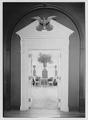 The image depicts a black and white photograph of an elegant room, likely part of a grand estate or mansion. The focal point is the entryway which features a tall, ornate arch with classical design elements such as columns on either side and decorative moldings around its edges. Atop the arch sits what appears to be a bird-like sculpture.

Through this arched entrance, we can see into another room that serves as a sunroom or conservatory. The interior of the next space is well-lit by natural light streaming in through large windows or glass doors on either side and at one end. In contrast with the darker tones outside, the inside appears brighter, creating an inviting atmosphere.

Within this sunroom setting, there's a prominent centerpiece: a vase filled with flowers placed centrally on what looks like a table draped with a cloth. The arrangement of furniture includes chairs arranged around another piece of decorative fabric or possibly curtains that frame the space elegantly. This seating area is flanked by two identical vases containing plants or sculptures.

The overall impression conveyed through this photograph suggests an atmosphere of sophistication and elegance, reflective of its likely historical context during the late 19th century to early 20th century when such architectural features were popular in residential design.