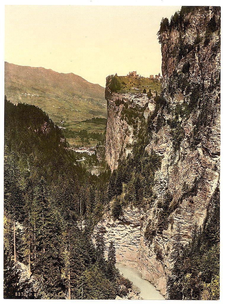 The image depicts a stunning mountainous landscape, possibly captured during the late 19th century. Dominating the scene is a steep cliff with rugged terrain and sparse vegetation clinging to its surface. The rocky face of the cliff transitions into lush greenery at higher elevations, suggesting varied altitudes within this natural setting.

Below the towering cliff, a serene river snakes through the valley floor, reflecting the muted tones of its surroundings. Its banks are lined with dense forestation that adds contrast against the greyish hue of the water and the earthen colors above. The greenery extends towards rolling hills in the background, where patches of cultivated land hint at human habitation.

Further back on these gentle slopes stands a small village or settlement composed of several buildings clustered together, nestled within this bucolic setting. Above it all lies an expansive view of more mountains fading into the horizon under a soft sky with light cloud cover.

This picturesque scene is reminiscent of early color photography techniques used during that era for artistic purposes such as postcards and album images to capture the beauty of nature or scenic views, likely intended to promote tourism in regions like Grisons, Switzerland. The warm sepia tones give it an aged look, evoking a sense of nostalgia while showcasing the timeless allure of this Alpi [...]