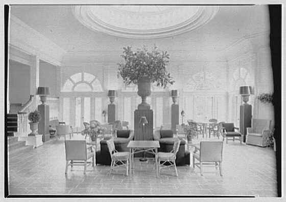 The image depicts a spacious sunroom with high ceilings and large windows, allowing ample natural light to illuminate the space. In the center stands a tall vase filled with lush greenery, serving as an elegant focal point. Surrounding this vase are several white chairs paired with small side tables, creating a cozy seating arrangement. The flooring appears to be polished wood or tile, reflecting some of the sunlight and adding warmth to the ambiance.

Lining one wall is a series of potted plants that enhance the room's connection to nature. On either side of the sunroom are additional pieces of furniture: sofas on the left and armchairs with matching tables on the right, all arranged symmetrically. The color palette seems monochromatic in this black-and-white photograph, emphasizing shades of gray, white, and green.

The architecture includes classical features such as arched windows at the far end, contributing to a sense of grandeur and openness within the space. Overall, the sunroom exudes an atmosphere of sophistication and tranquility, inviting relaxation or conversation amidst its elegant setting.
