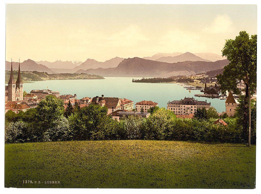 The image depicts a picturesque landscape featuring a large lake in the foreground, with a range of mountains forming a majestic backdrop. The shoreline is lined with an array of buildings and structures indicative of European architecture, including what appears to be churches or historical edifices on one side, possibly indicating a town or cityscape beside the water's edge.

The colors are vivid yet have a slightly faded quality typical of aged postcards or photographs from the late 19th century. The vegetation is lush and green with patches of white flowers adding contrast against the verdant foliage in the foreground grassland area that extends into the middle ground, where clusters of trees can be seen.

The overall composition suggests this image captures a serene moment in Lucerne, Switzerland during what seems to have been late spring or summer. The inclusion of text at the bottom indicates it is likely an old postcard with historical significance, possibly taken from a vantage point overlooking the lake and town on a hillside or elevated ground.

The reference provided states that this image falls within the timeframe where such photochrom prints were popular for travel memorabilia during that era.