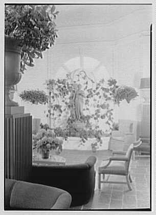 The image depicts a sunroom with an ornate fountain as its centerpiece, surrounded by potted plants and seating arrangements. The room appears to be well-lit from natural light coming through the windows or glass panels. There are several chairs and benches placed around the space for relaxation or socializing.
Additional information about this specific photo can be found in "The Homestead" publication, which is associated with Hot Springs, Virginia dated May 15th of an unspecified year (likely early to mid-20th century based on style). The image credits Gottschol-Schleisner Inc., a company that produced photographic negatives. This particular negative measures 5x7 inches in size and has the identifier "691e" with its corresponding reference code "691efd13450e57a2674fee21".
