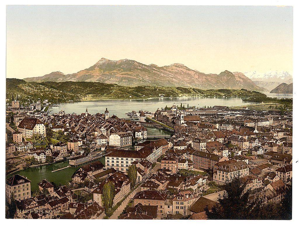 The image is a vintage photograph depicting an expansive and picturesque view over the cityscape of Lucerne, Switzerland. The perspective appears to be from a high vantage point overlooking numerous buildings with steeply pitched roofs characteristic of European architecture. A river winds through the urban area, reflecting sunlight on its surface.

In the distance, calm waters extend towards mountains that rise majestically against a clear sky. The water body is likely Lake Lucerne or another significant lake in Switzerland, surrounded by hills and mountainous regions showcasing various shades of green vegetation with patches of brown indicating forested areas.

The image has an aged appearance typical of early 20th-century photographs, suggesting it was taken around the late 1890s to early 1900s. The quality and style indicate that this could be a photographic print from glass negatives or film common in photochrom printing during those years. Notable features include the detailed rendering of buildings with varying architectural styles, indicating both residential and commercial areas.

The photograph is part of the collection "Views of Switzerland" by From Gutsch, Lucerne, as referenced on the image file's source link provided. This context implies that it was likely produced for tourism or illustrative purposes during a period when Swiss scenery attracted attention d [...]