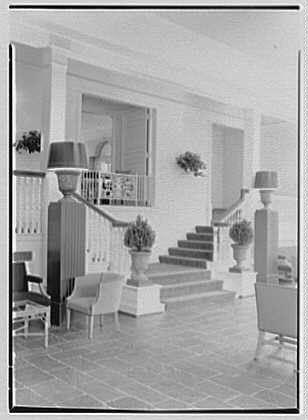 The image is a black and white photograph of an elegant sunroom, located at The Homestead in Hot Springs, Virginia. It appears to be from May 15th, 1946. This particular photo captures the interior details of the stairway leading up to what seems like another level or section within this building.
The room is adorned with a set of stairs that feature large urn-shaped planters at their corners and on top, which hold potted plants. The steps themselves are bordered by low railings for safety. The flooring consists of flat stone tiles laid out in an orderly pattern throughout the space. On either side of the staircase, there's seating furniture: a couple of armchairs with slender legs stand prominently near each stairwell.
The architecture and design suggest that this setting is part of a historical or upscale establishment. It reflects a classic interior style from mid-20th-century America where comfort was combined with an appreciation for natural materials like stone in the decor choices. The lighting appears to be artificial, as indicated by several large lamps placed around the room on pedestals and stands.
The image is credited to Gottscho-Schleisner Inc., which suggests that it's part of their collection or archive documenting architectural elements from various locations during this period. Overall, the scene conveys an atmosphere of tranquility and leisure associate [...]