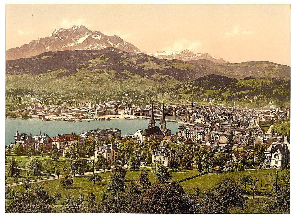 This image depicts a picturesque landscape featuring the cityscape and natural beauty of Lucerne, Switzerland. In the foreground is a well-maintained park with neatly trimmed trees and grassy areas providing green space amidst urban development. The middle ground reveals an array of historical buildings showcasing European architectural styles, including pointed roofs, ornate facades, and spire-topped structures that hint at their religious or civic significance.

In the background stands a prominent mountain range with snow-capped peaks dominating the skyline above Lucerne's urban setting. Below these mountains is a river flowing through the city, leading to an expansive lake dotted with boats in its calm waters. The overall scene captures a harmonious blend of nature and architecture, evoking the charm and historical allure of Switzerland during what appears to be late 19th-century Europe.

Additional information about this image can be found from Pilatus and Lucerne, seen from Drei Linden, Lucerne, Switzerland between ca. 1890 and ca. 1900; Views of Switzerland on the website images.loener.nl/PhotochromPrints/full/65e4/65e47a2a5890ae069f67c3ac.jpg