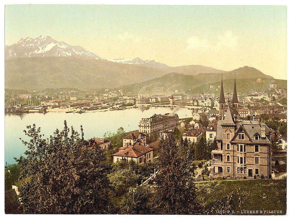 The image depicts a picturesque town nestled by the water, with mountains rising in the background. The architecture is European, featuring steeples and ornate buildings. A bridge spans across the river or lake that flows through the town. In the foreground are lush green trees framing the view of the quaint settlement below.