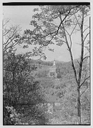 The image is a black and white photograph depicting a scenic view of The Homestead in Hot Springs, Virginia. In the distance, there's an imposing tower that stands out against the surrounding landscape. This historic structure appears to be elevated on stilts or pillars over a body of water, which adds to its grandeur.

The foreground features trees with bare branches suggesting it might be during late autumn or early winter months. The perspective from behind foliage creates a sense of mystery and draws attention toward the tower as the focal point of the image. This particular photograph was taken on May 15th in 1946, showcasing its historical context.

The caption "Tower from distance" provides valuable information about what is being depicted, while additional details found in The Homestead resource offer deeper insights into this iconic American landmark and its significance at that time period.