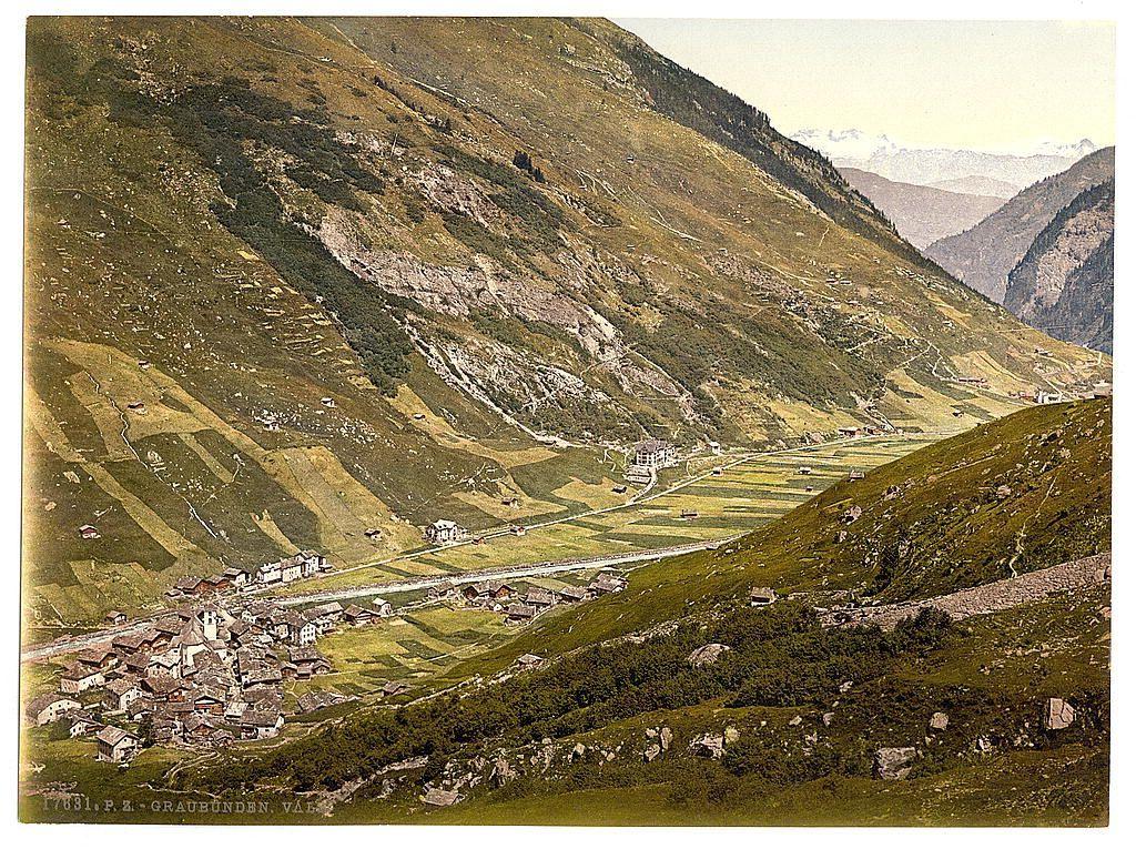 The image depicts a picturesque, expansive view of Graubünden Valley in Vals. It showcases steep mountains adorned with patches of greenery and brown earth tones, indicative of either early spring or late fall. A village nestled at the valley's base is visible; it features traditional architecture with dark roofs contrasted against lighter walls. The buildings appear closely packed together along a central street that winds through them. Scattered across the landscape are terraced fields in shades of green and brown, suggesting agricultural activity. In the distance, faint outlines hint at additional mountainous terrain or valleys beyond. This scene is rendered as an artistic print with text indicating it was created between 1890 and 1900 by P.E.Graubunden, Valais. The overall atmosphere of the image radiates tranquility and rustic charm reflective of Swiss countryside life in that era.