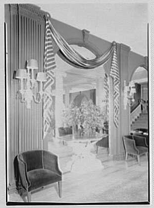 The image is a black and white photograph of an elegant room with classic decor, likely taken in the mid-20th century. The architecture features ornate columns draped with fabric or drapery, adding to the opulent atmosphere.

In the foreground, there are two chairs: one dark-colored armchair on the left side facing away from the camera and another chair further back positioned parallel to a wooden floor that extends into the distance. The seating arrangement suggests an inviting space for social gatherings or relaxation within this grand setting.

The main focus of the room appears to be a hallway leading towards additional areas, with potted plants visible in the background on either side of it. This gives off a sense of depth and continuity as you look further back into the room's interior architecture.

Overall, the photograph captures an ambiance that reflects sophistication, comfort, and attention to detail typical of historic ballrooms or elegant public spaces during this time period.
